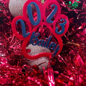May include: Red paw print Christmas ornament with the year 2023 and the name Bailey in blue lettering.