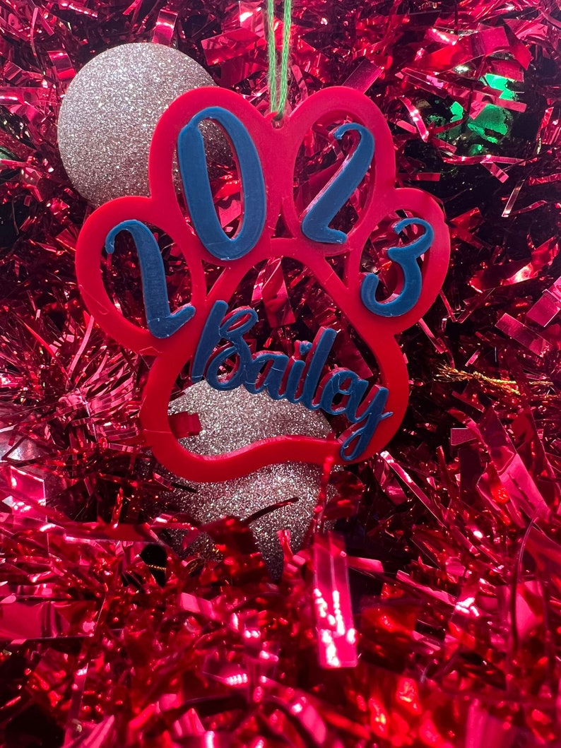 May include: Red paw print Christmas ornament with the year 2023 and the name Bailey in blue lettering.