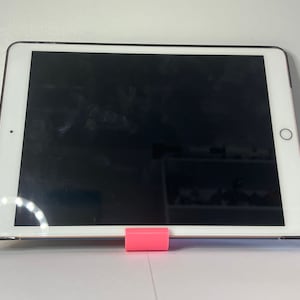 May include: A white tablet computer with a black screen is propped up on a pink stand. The tablet is on a white surface.
