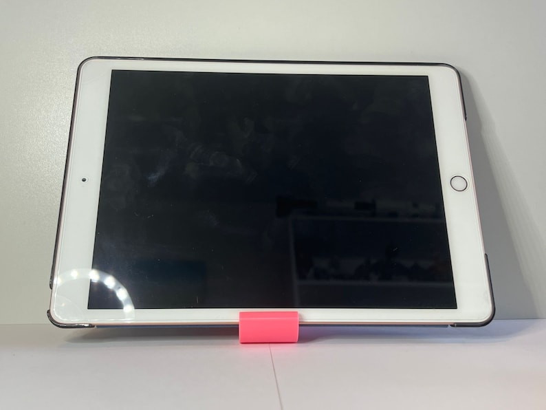 May include: A white tablet computer with a black screen is propped up on a pink stand. The tablet is on a white surface.