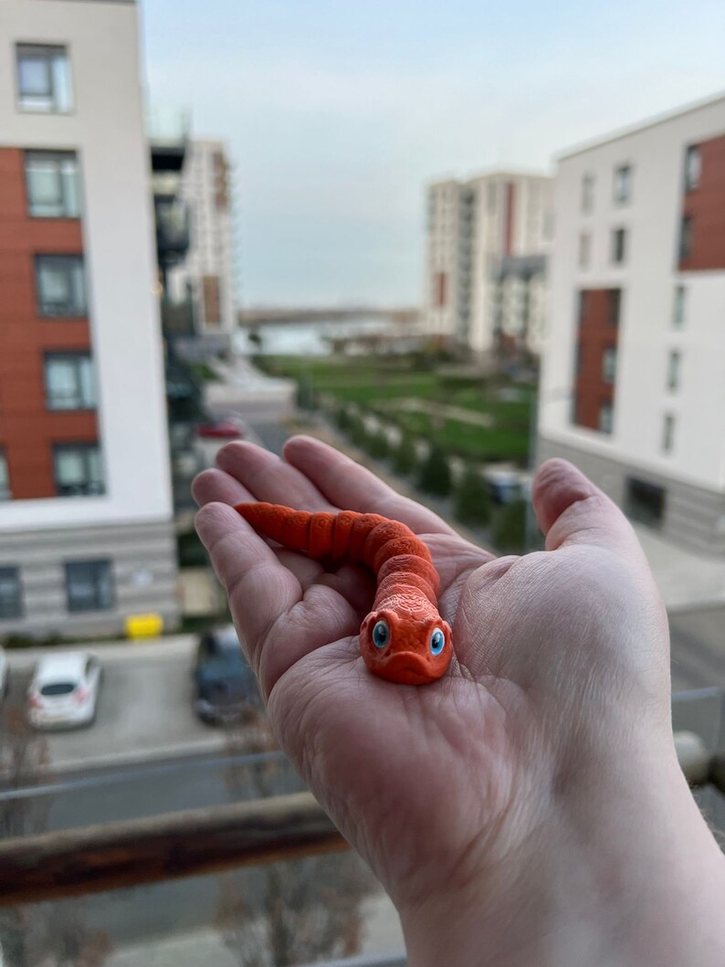 May include: A hand holding a bright orange worm toy with blue eyes. The toy is made of a soft, pliable material.