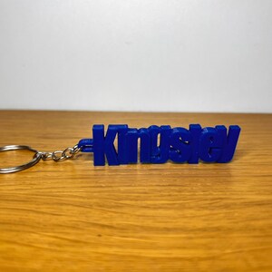 Op de afbeelding: Een blauwe plastic sleutelhanger met de naam "Kingsley" erop gedrukt.