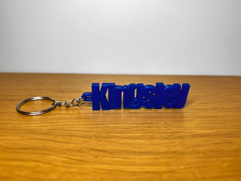 Op de afbeelding: Een blauwe plastic sleutelhanger met de naam "Kingsley" erop gedrukt.