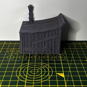 May include: A gray, wooden miniature building with a chimney and a roof. The building is sitting on a green cutting mat with yellow markings.