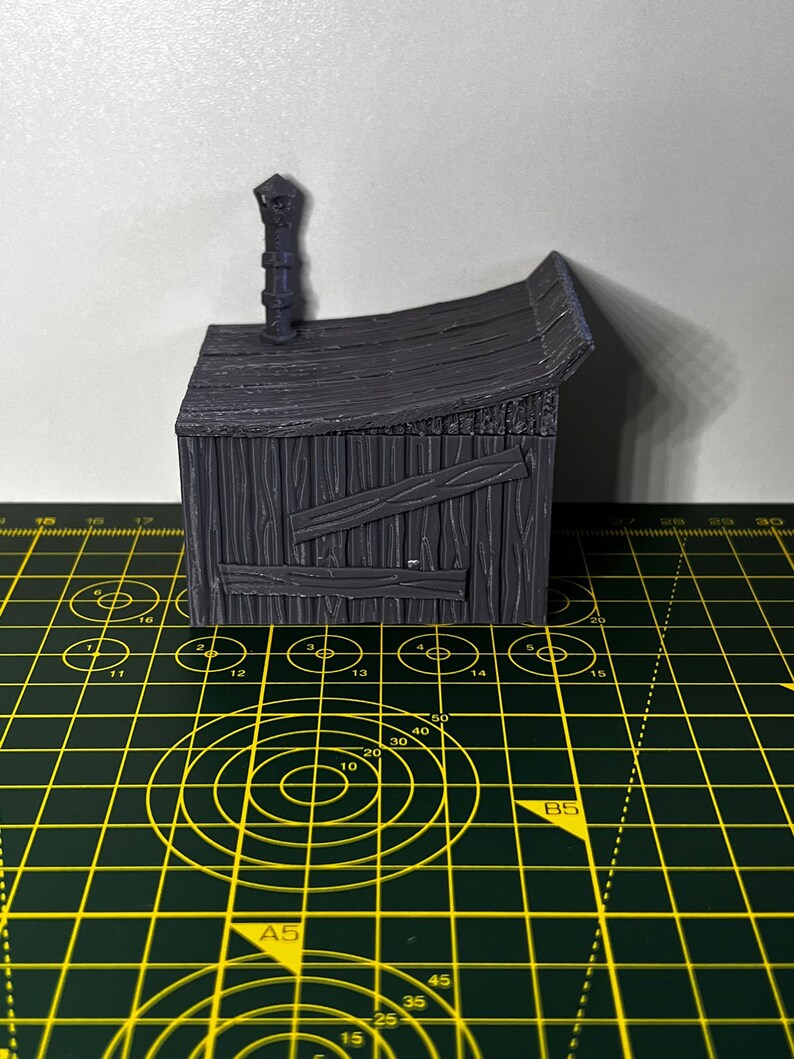 May include: A gray, wooden miniature building with a chimney and a roof. The building is sitting on a green cutting mat with yellow markings.