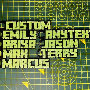 Może przedstawiać: Czarne i zielone niestandardowe breloczki do kluczy z imionami i słowem "CUSTOM" w pikselowanej czcionce. Imiona to "EMILY", "ANYTEXT", "ARIYA", "JASON", "MAX", "TERRY" i "MARCUS". Każdy breloczek ma okrągły otw&oacute;r.