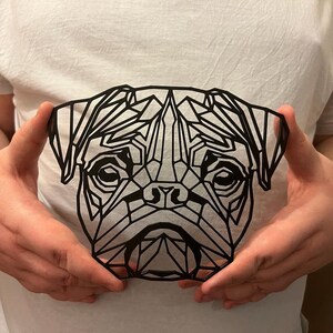 May include: A black geometric design of a pug's face, cut out of paper or cardstock, being held by a person's hands.