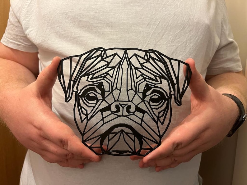 May include: A black geometric design of a pug's face, cut out of paper or cardstock, being held by a person's hands.