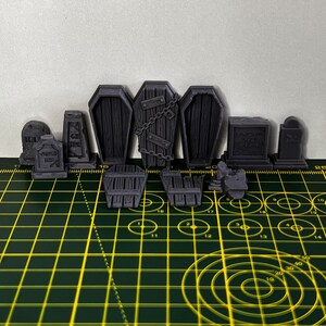 May include: A set of 3D printed Halloween decorations, including tombstones, coffins, and gates. The decorations are painted in a dark grey colour and have a textured surface. The decorations are arranged on a green cutting mat with yellow grid lines.