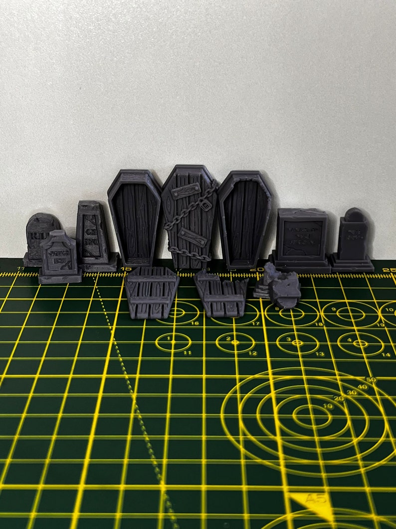May include: A set of 3D printed Halloween decorations, including tombstones, coffins, and gates. The decorations are painted in a dark grey colour and have a textured surface. The decorations are arranged on a green cutting mat with yellow grid lines.