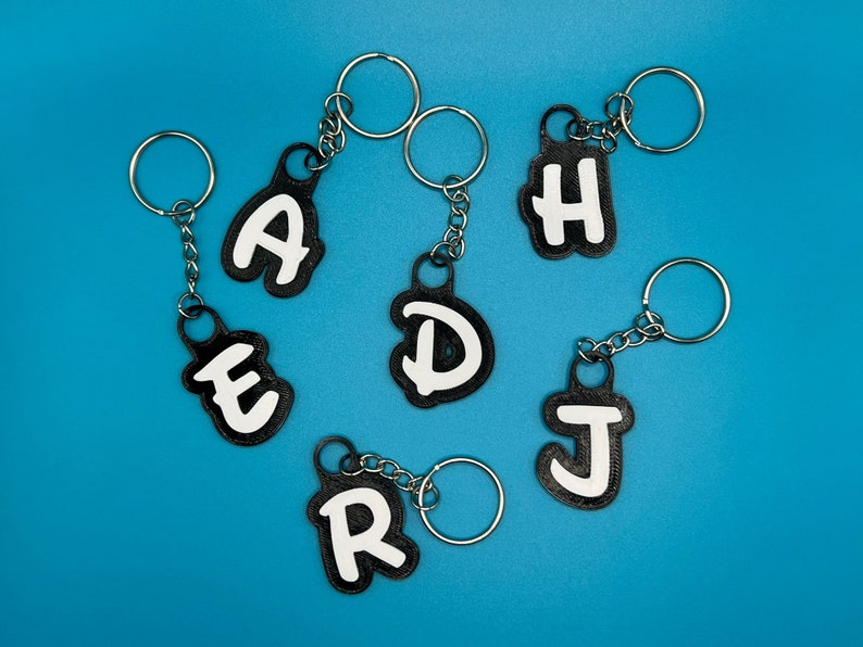 Op de afbeelding: Zes zwarte sleutelhangers met witte letters die het woord "RADJED" spellen op een blauwe achtergrond.