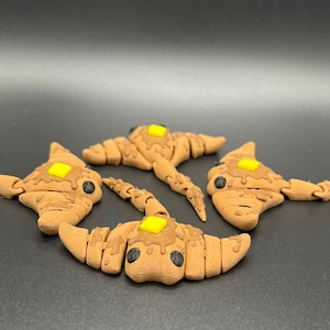 Puede incluir: Cuatro juguetes con forma de croissant impresos en 3D de color marrón y amarillo con ojos negros y un cuadrado amarillo en la parte superior.