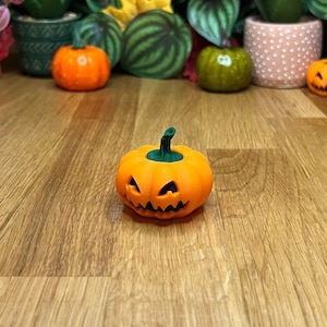 Puede incluir: Una pequeña calabaza naranja con una cara de calabaza tallada. La calabaza tiene un tallo verde y está sentada sobre una superficie de madera.