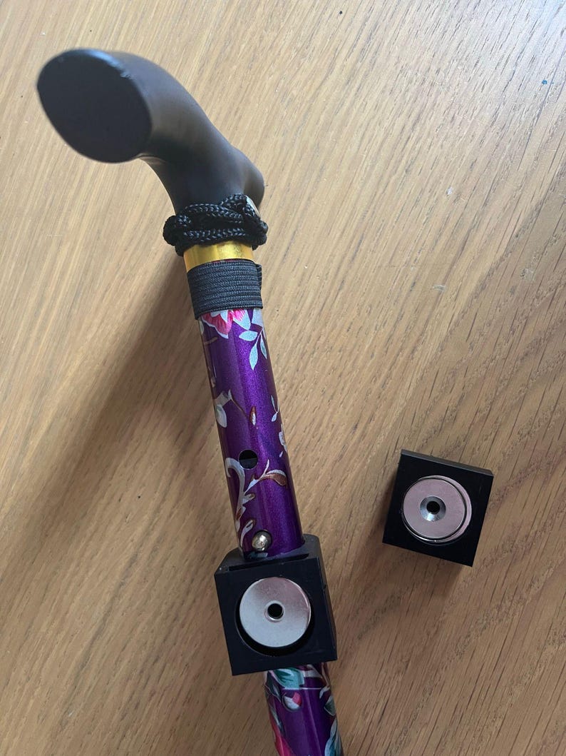 May include: A purple walking stick with a floral pattern and a black handle. The stick has a black magnetic attachment with a silver center. There is a second black magnetic attachment with a silver center on the table.