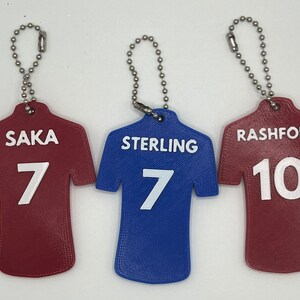May include: Three red and blue jersey-shaped keychains with white lettering. The keychains feature the names "Saka 7", "Sterling 7", and "Rashford 10".