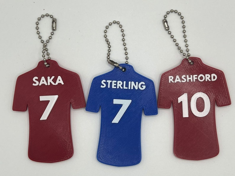 May include: Three red and blue jersey-shaped keychains with white lettering. The keychains feature the names "Saka 7", "Sterling 7", and "Rashford 10".