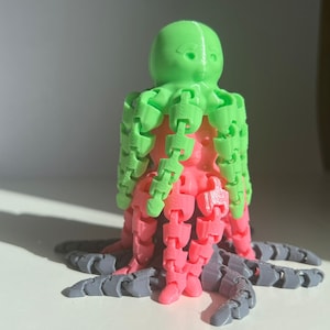以下が含まれることがあります： セグメントされた腕を持つ3Dプリントされたタコのおもちゃ。頭は緑色、上腕はピンク色、下腕は灰色です。