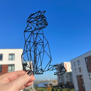 May include: A black geometric cat sculpture made of wire. The sculpture is held in a hand against a blue sky background.