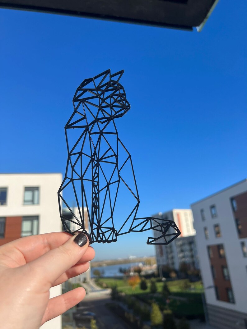 May include: A black geometric cat sculpture made of wire. The sculpture is held in a hand against a blue sky background.
