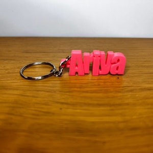 Op de afbeelding: Roze plastic sleutelhanger met het woord "Ariva" gespeld in blokletters.