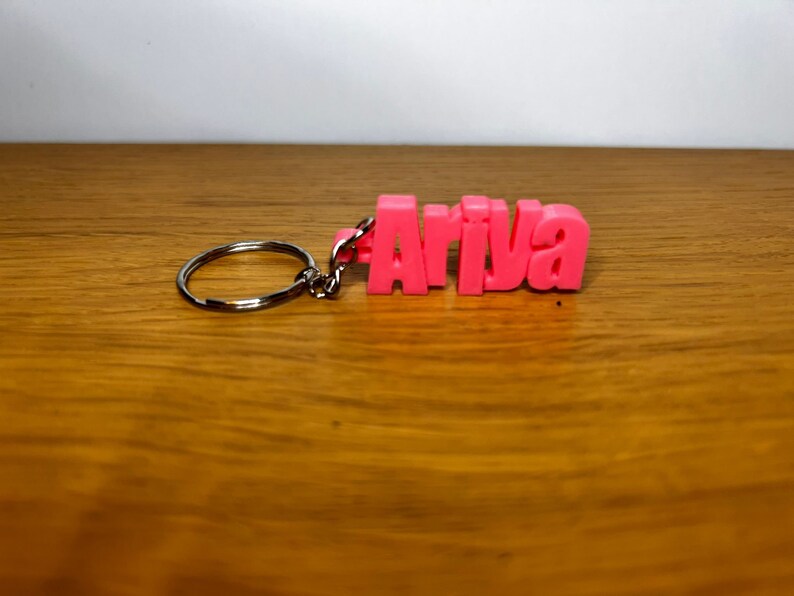 Op de afbeelding: Roze plastic sleutelhanger met het woord "Ariva" gespeld in blokletters.