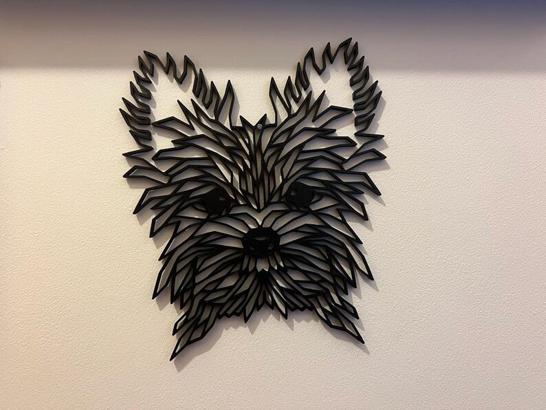 May include: Black and white geometric silhouette of a Yorkshire Terrier dog's head, made of layered paper or wood, mounted on a white wall.