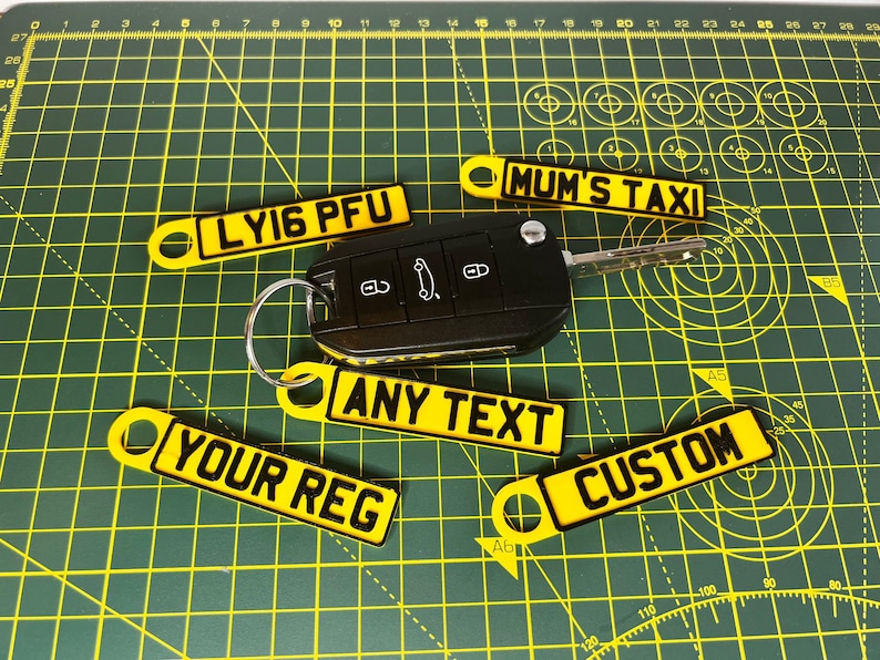 May include: Five yellow keychains with black text. The keychains are shaped like license plates and have the following text: "LYI6 PFU", "MUM'S TAXI", "ANY TEXT", "YOUR REG", and "CUSTOM".
