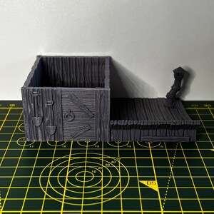 May include: A gray 3D printed miniature wooden building with a hinged door and a small lamppost on the side. The building has a textured wood grain design and a small opening in the front.