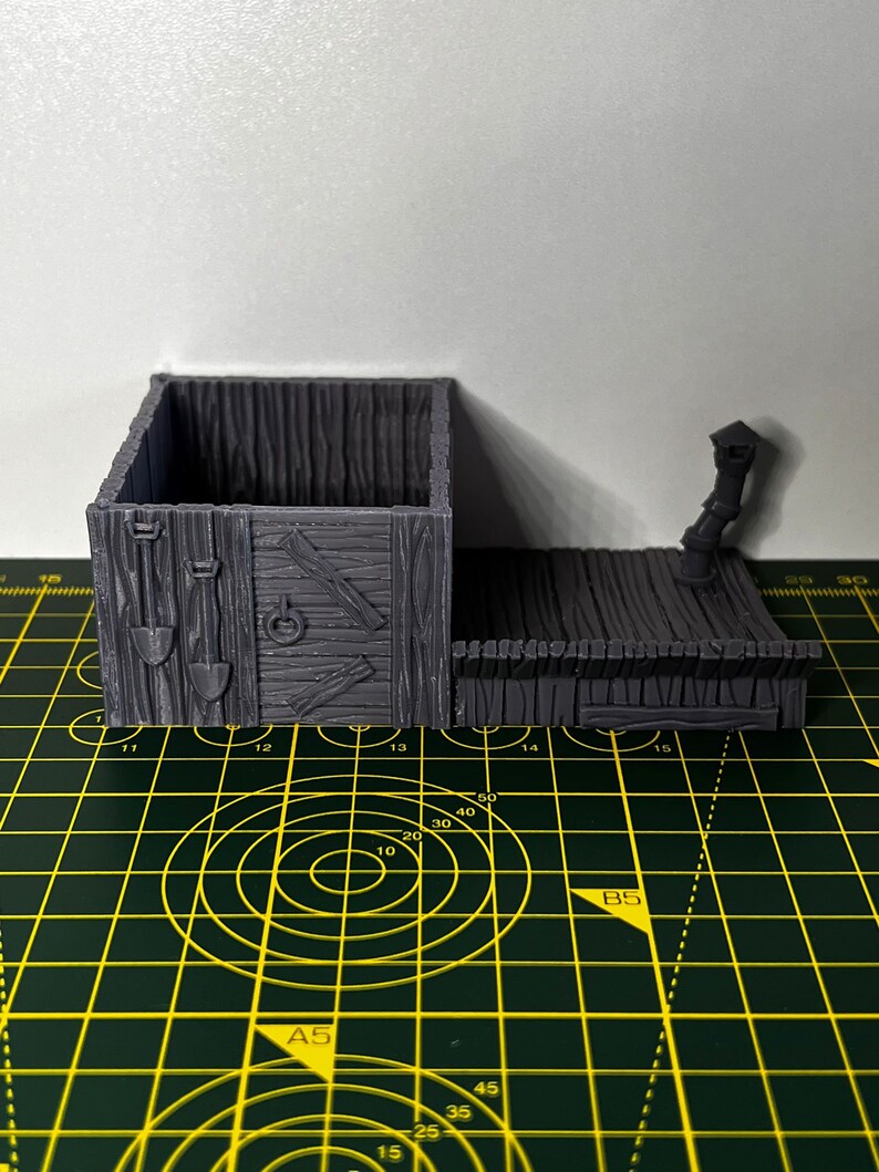 May include: A gray 3D printed miniature wooden building with a hinged door and a small lamppost on the side. The building has a textured wood grain design and a small opening in the front.
