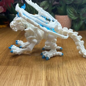 Puede incluir: Un esqueleto de drag&oacute;n de pl&aacute;stico blanco con detalles azules. El drag&oacute;n est&aacute; en posici&oacute;n sentada con las alas extendidas.