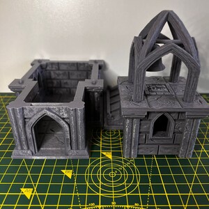 May include: Two gray 3D printed miniature buildings with gothic arches, one with a bell tower. The buildings are on a green cutting mat with yellow grid lines.
