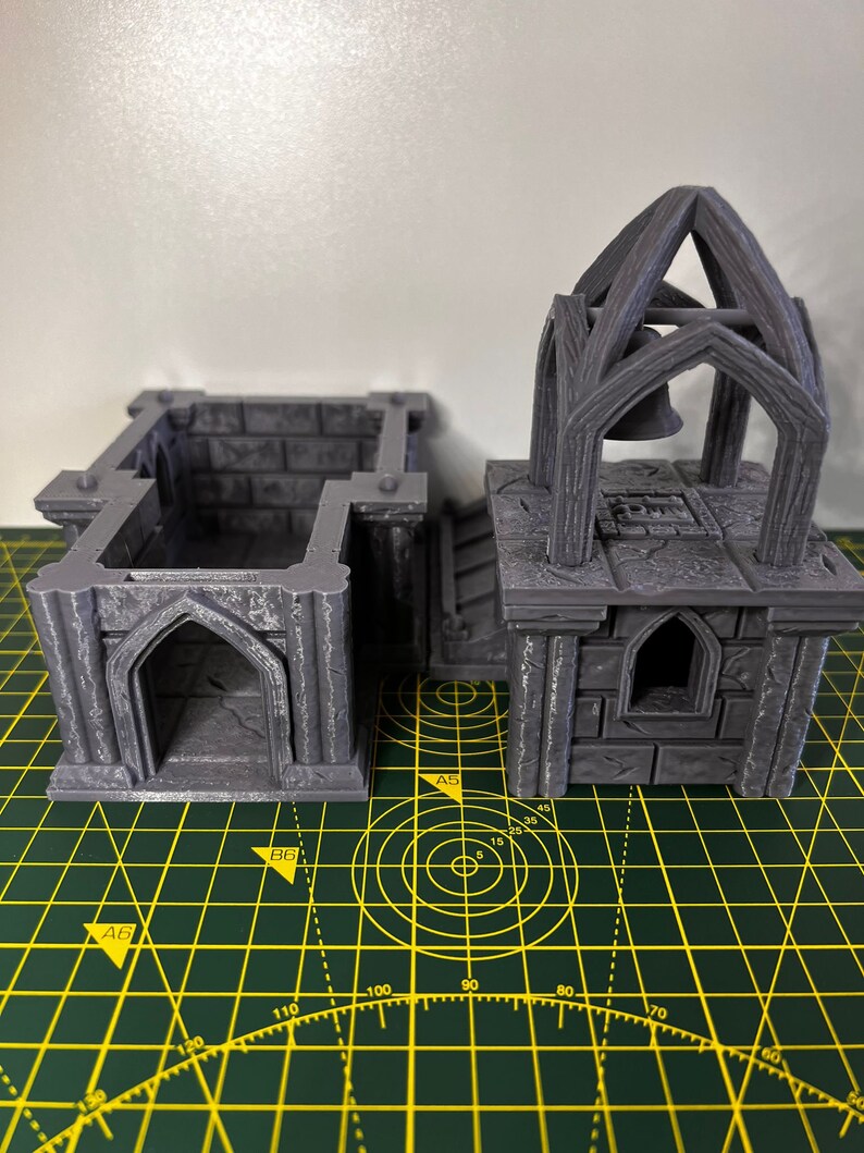 May include: Two gray 3D printed miniature buildings with gothic arches, one with a bell tower. The buildings are on a green cutting mat with yellow grid lines.