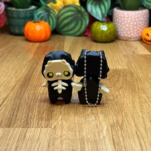 Puede incluir: Dos figuras con temática de Halloween. Una es un esqueleto negro con caja torácica y brazos blancos. La otra es un gato negro con una cadena plateada. Las figuras están sobre una superficie de madera con calabazas y plantas al fondo.