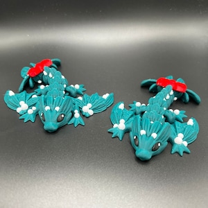May include: Two teal green, 3D printed, lizard-like figurines with white spots and red accents. The figurines have wings and are posed on a black surface.