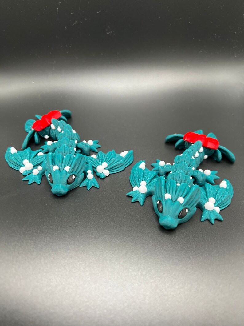 May include: Two teal green, 3D printed, lizard-like figurines with white spots and red accents. The figurines have wings and are posed on a black surface.