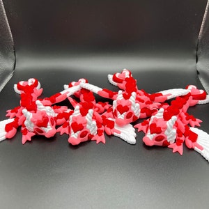 Puede incluir: Cinco peque&ntilde;as figuras de drag&oacute;n de color rosa y blanco con escamas en forma de coraz&oacute;n. Los dragones tienen alas y est&aacute;n acostados sobre una superficie negra.