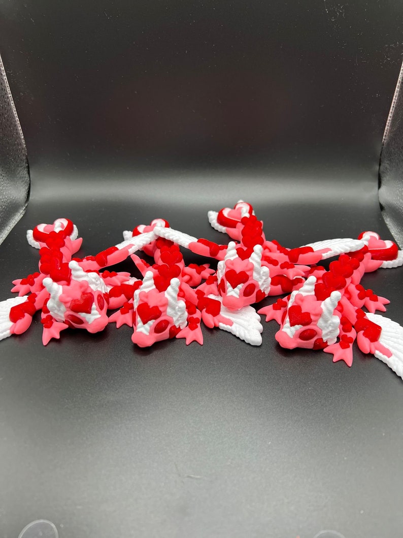 Puede incluir: Cinco peque&ntilde;as figuras de drag&oacute;n de color rosa y blanco con escamas en forma de coraz&oacute;n. Los dragones tienen alas y est&aacute;n acostados sobre una superficie negra.