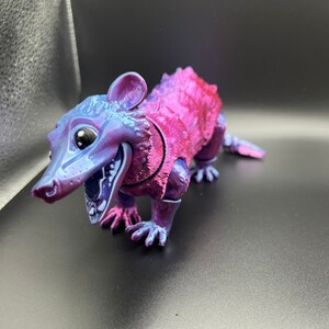 May include: A small, purple and pink plastic toy opossum with a wide open mouth and large eyes. The toy is posed in a standing position with its front legs extended.