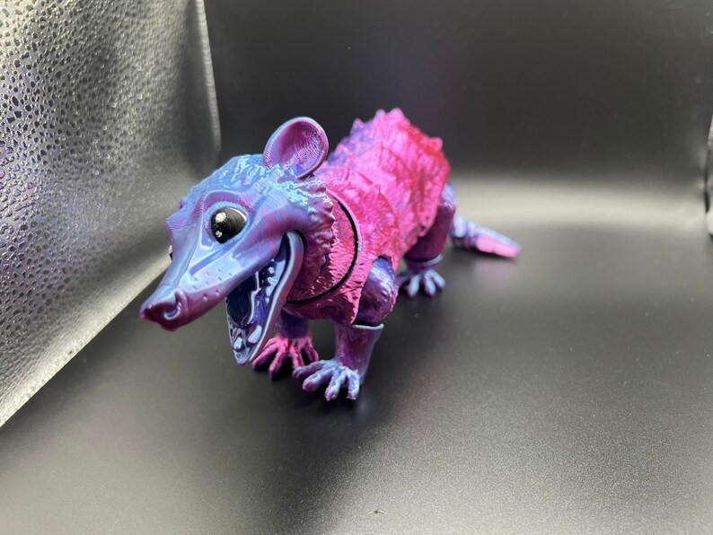 May include: A small, purple and pink plastic toy opossum with a wide open mouth and large eyes. The toy is posed in a standing position with its front legs extended.