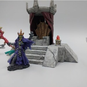 May include: A miniature fantasy scene with a green-skinned figure wearing a purple robe and a crown, holding a red staff. The figure stands on a stone platform with a throne and red curtains in the background. Two other figures are visible in the background.