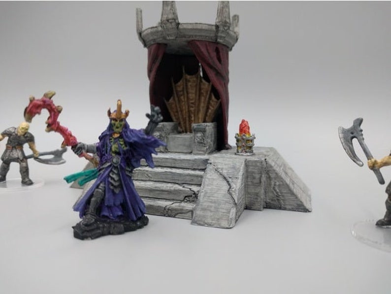 May include: A miniature fantasy scene with a green-skinned figure wearing a purple robe and a crown, holding a red staff. The figure stands on a stone platform with a throne and red curtains in the background. Two other figures are visible in the background.