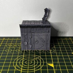 May include: A gray 3D printed miniature of a small wooden building with a chimney and a door. The building has a textured wooden surface and a roof with a slight overhang. The miniature is sitting on a green and yellow cutting mat.