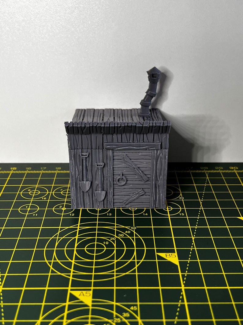 May include: A gray 3D printed miniature of a small wooden building with a chimney and a door. The building has a textured wooden surface and a roof with a slight overhang. The miniature is sitting on a green and yellow cutting mat.
