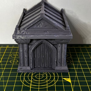 May include: A grey, detailed miniature mausoleum with a gabled roof and a closed door. The mausoleum is sitting on a green cutting mat with yellow markings.