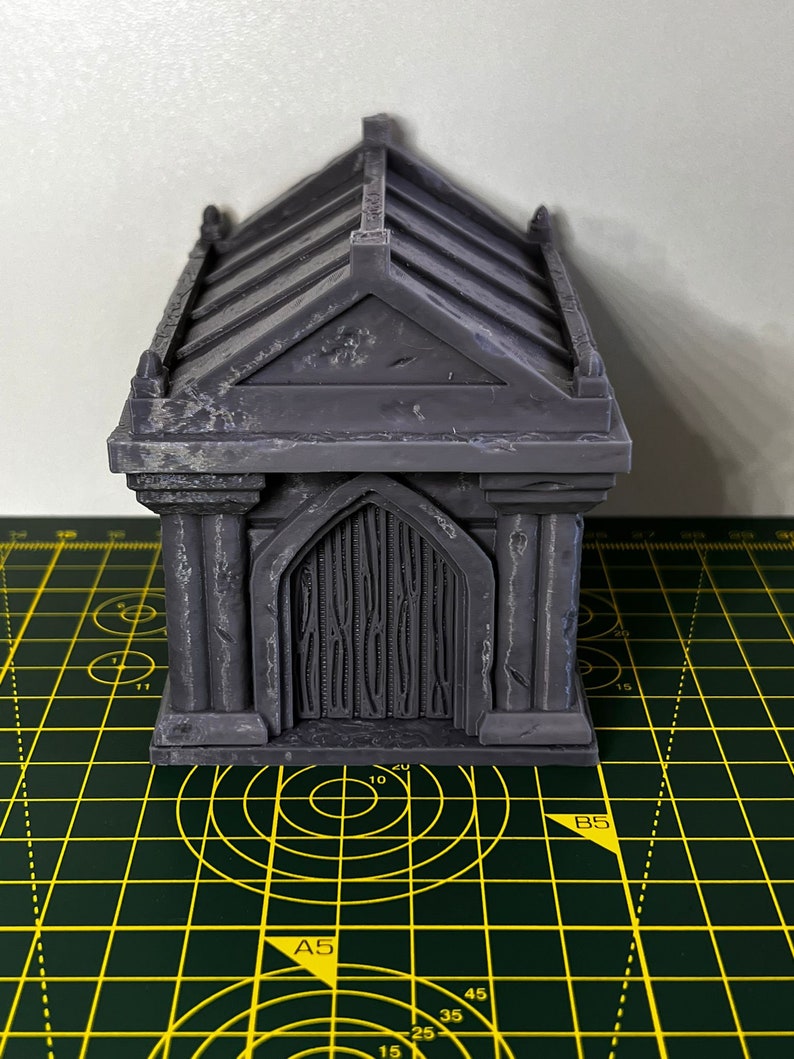 May include: A grey, detailed miniature mausoleum with a gabled roof and a closed door. The mausoleum is sitting on a green cutting mat with yellow markings.