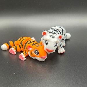 May include: Two small tiger figurines, one orange and black, the other white with black stripes. The orange tiger is lying down with its head turned to the side, while the white tiger is standing with its head tilted down. Both tigers have black eyes and pink noses.