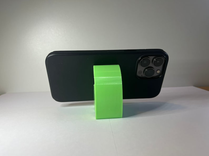 May include: A black smartphone with a green phone stand. The phone is in a black case and the stand is made of plastic.
