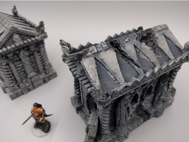 May include: Two detailed, gray, stone miniature buildings with a weathered, aged look. The buildings are in the style of a mausoleum or crypt. One building has a broken roof.