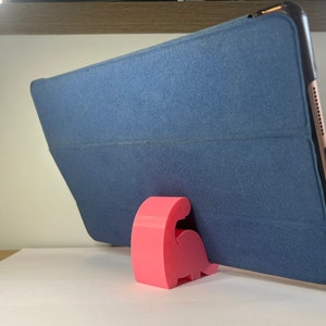 May include: A pink dinosaur-shaped phone stand holds a blue tablet case in a vertical position.