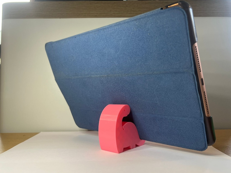 May include: A pink dinosaur-shaped phone stand holds a blue tablet case in a vertical position.